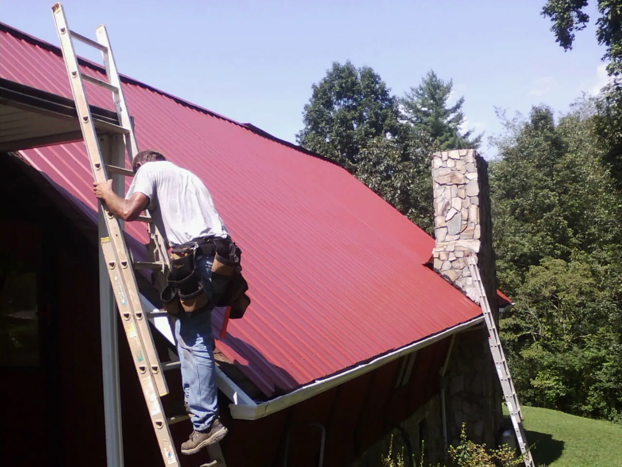 Roofer on a ladder beside a red metal roof and stone chimney — Soffit & Fascia Repair by Roofing Brothers in Valley Park