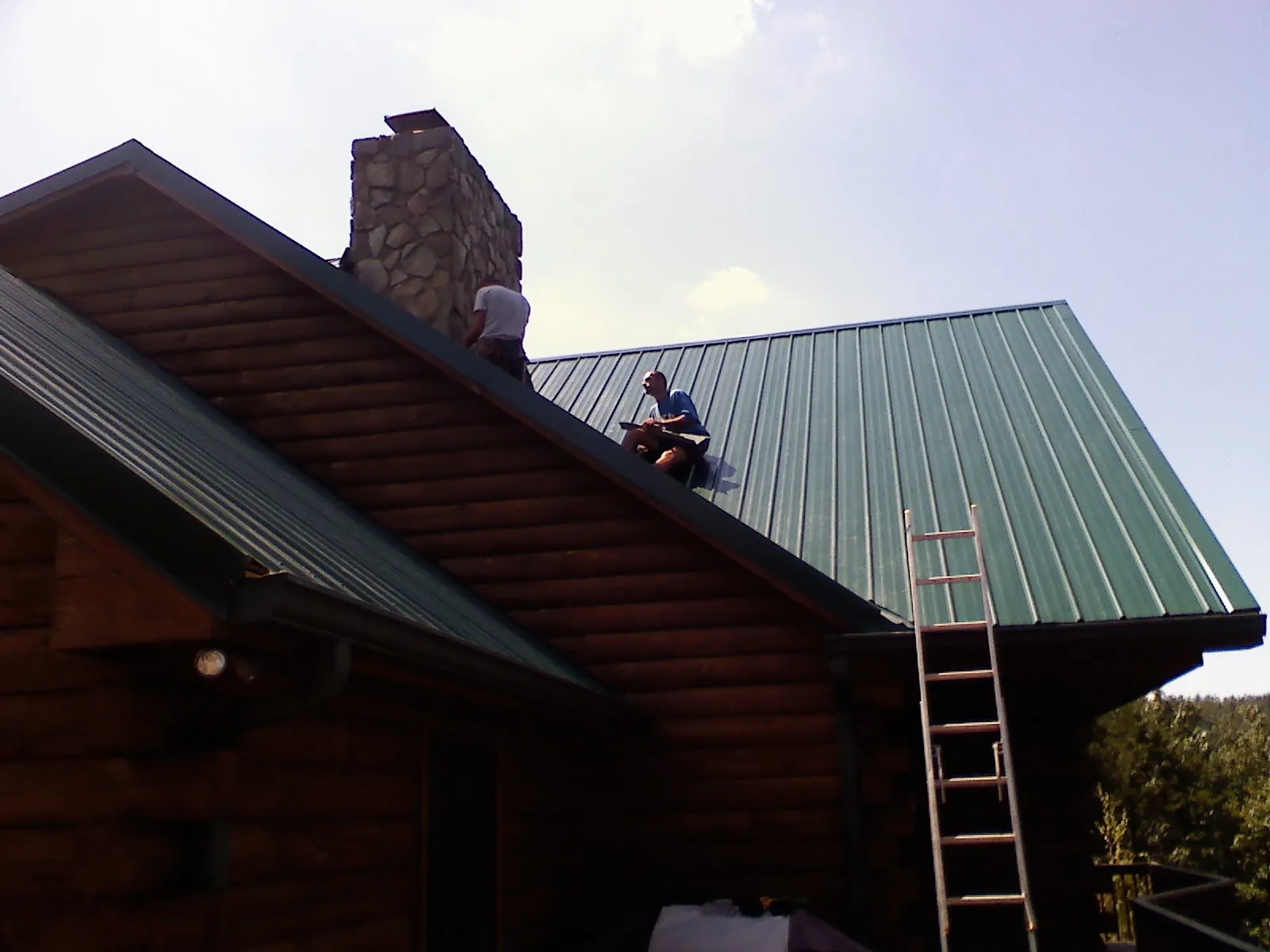 Roofers installing green metal panels beside a stone chimney on a log home in Valley Park